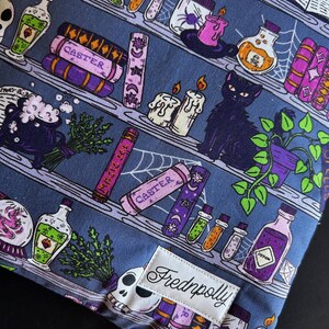 May include: A fabric item with a repeating pattern of shelves filled with potion bottles, spell books, candles, and a black cat. The color scheme includes purple, green, and white elements on a blue background. The label reads "Fredpolly".
