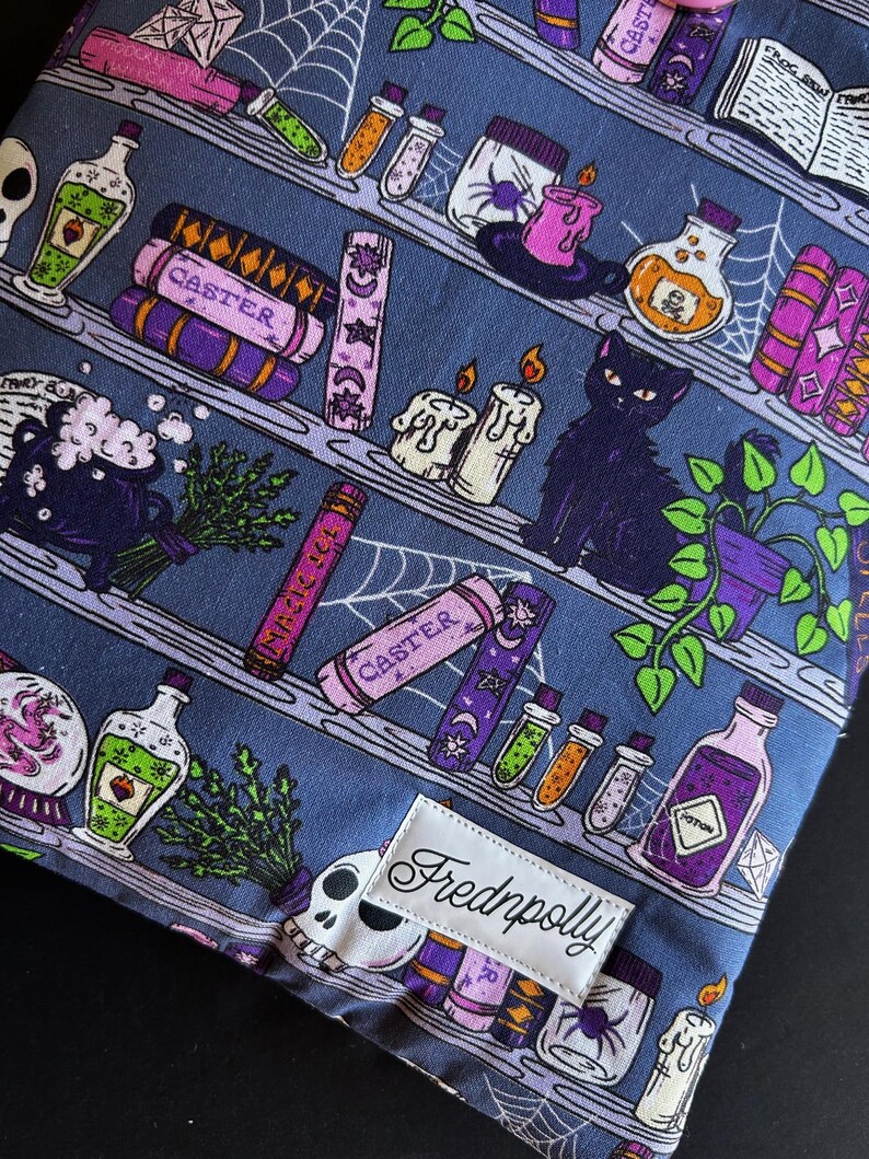 May include: A fabric item with a repeating pattern of shelves filled with potion bottles, spell books, candles, and a black cat. The color scheme includes purple, green, and white elements on a blue background. The label reads "Fredpolly".