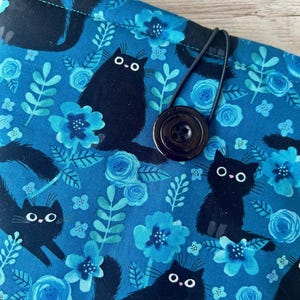 Puede incluir: Una bolsa de tela azul con cierre de bot&oacute;n negro y una correa el&aacute;stica negra. La tela presenta un patr&oacute;n repetitivo de gatos negros y flores azules.