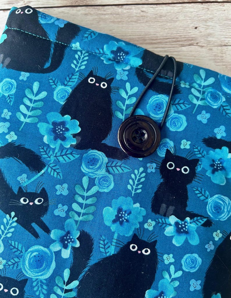 Puede incluir: Una bolsa de tela azul con cierre de bot&oacute;n negro y una correa el&aacute;stica negra. La tela presenta un patr&oacute;n repetitivo de gatos negros y flores azules.