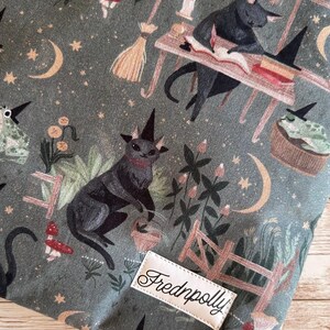May include: A fabric with a whimsical print featuring black cats wearing witch hats, mushrooms, moons, stars, and a small wooden fence. The fabric has a green background and a label that says "Frednpolly".