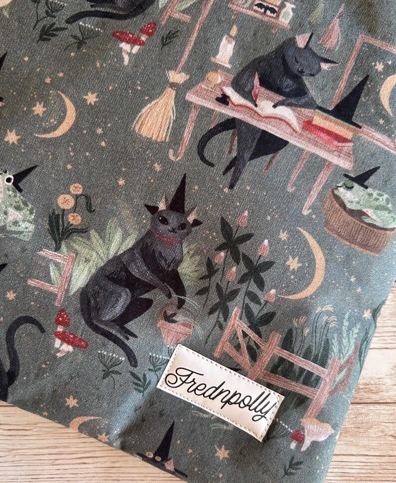 May include: A fabric with a whimsical print featuring black cats wearing witch hats, mushrooms, moons, stars, and a small wooden fence. The fabric has a green background and a label that says "Frednpolly".