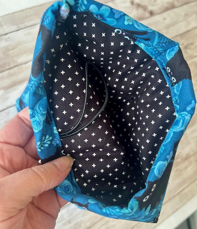 Puede incluir: Una bolsa de tela azul con un forro estampado en blanco y negro. La bolsa tiene un cord&oacute;n de ajuste negro y presenta un dise&ntilde;o de gato negro.