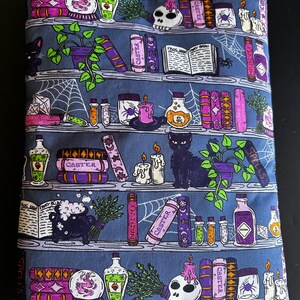 May include: A blue fabric book sleeve featuring a repeating pattern of bookshelves with potion bottles, spell books, candles, and a black cat. The word "CASTER" is printed on some books. A spider web design is also present.