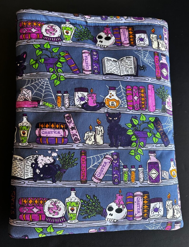 May include: A blue fabric book sleeve featuring a repeating pattern of bookshelves with potion bottles, spell books, candles, and a black cat. The word "CASTER" is printed on some books. A spider web design is also present.