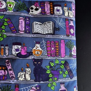 May include: A blue fabric with a repeating pattern of a witch's bookshelf. The design includes potion bottles, candles, books, a black cat, and a spiderweb. The books have the word "Caster" on them.