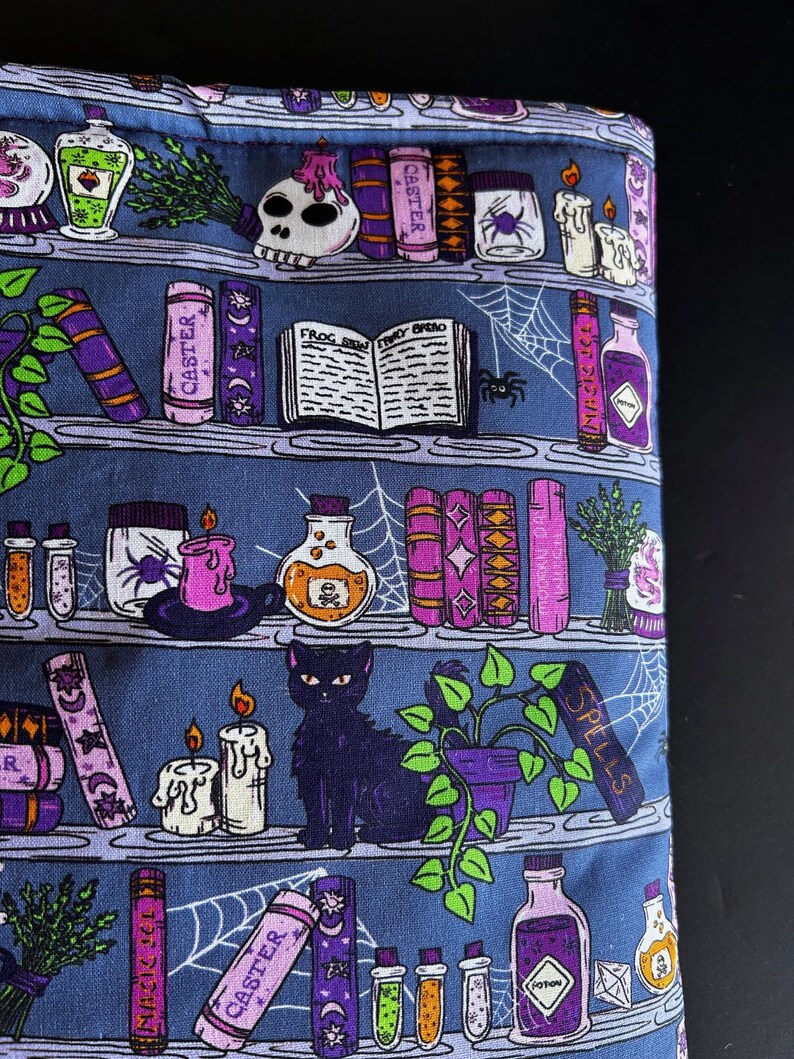 May include: A blue fabric with a repeating pattern of a witch's bookshelf. The design includes potion bottles, candles, books, a black cat, and a spiderweb. The books have the word "Caster" on them.