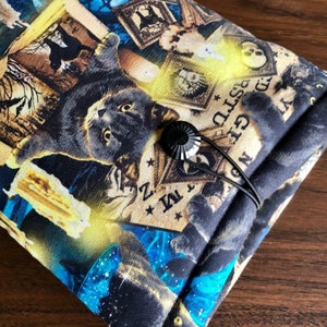 Op de afbeelding: Zwarte stoffen tas met een zwarte kat en verschillende vintage illustraties, waaronder kaarsen, schedels en een Ouija-bord. De tas heeft een zwarte koordsluiting met een zwarte knoop.