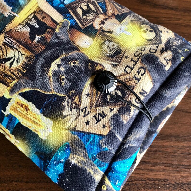 Op de afbeelding: Zwarte stoffen tas met een zwarte kat en verschillende vintage illustraties, waaronder kaarsen, schedels en een Ouija-bord. De tas heeft een zwarte koordsluiting met een zwarte knoop.