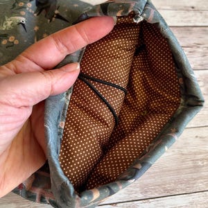 May include: A fabric pouch with a patterned exterior and a brown polka-dot interior. The exterior features a design with birds, plants, and stars in shades of blue, green, and pink. A black elastic cord is visible inside.