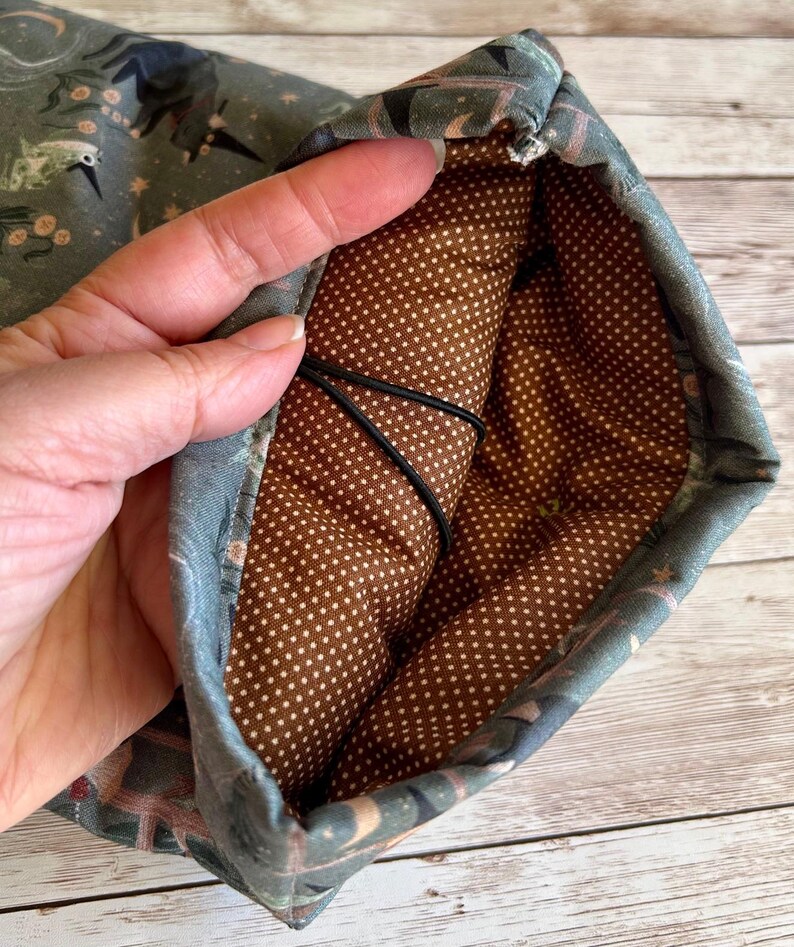 May include: A fabric pouch with a patterned exterior and a brown polka-dot interior. The exterior features a design with birds, plants, and stars in shades of blue, green, and pink. A black elastic cord is visible inside.