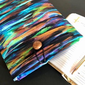 Könnte beinhalten: Ein Stoff-Notizbuch-Cover mit einem farbenfrohen abstrakten Design in Blau-, Grün-, Orange- und Schwarztönen. Der Umschlag wird mit einem braunen Knopf und einer schwarzen elastischen Schlaufe verschlossen. Ein teilweise sichtbares offenes Notizbuch und ein Stift sind im Hintergrund.