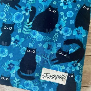 Puede incluir: Tela azul con un patr&oacute;n repetido de gatos negros y flores azules. La tela tiene una etiqueta blanca con el texto "Frednpolly" en ella.