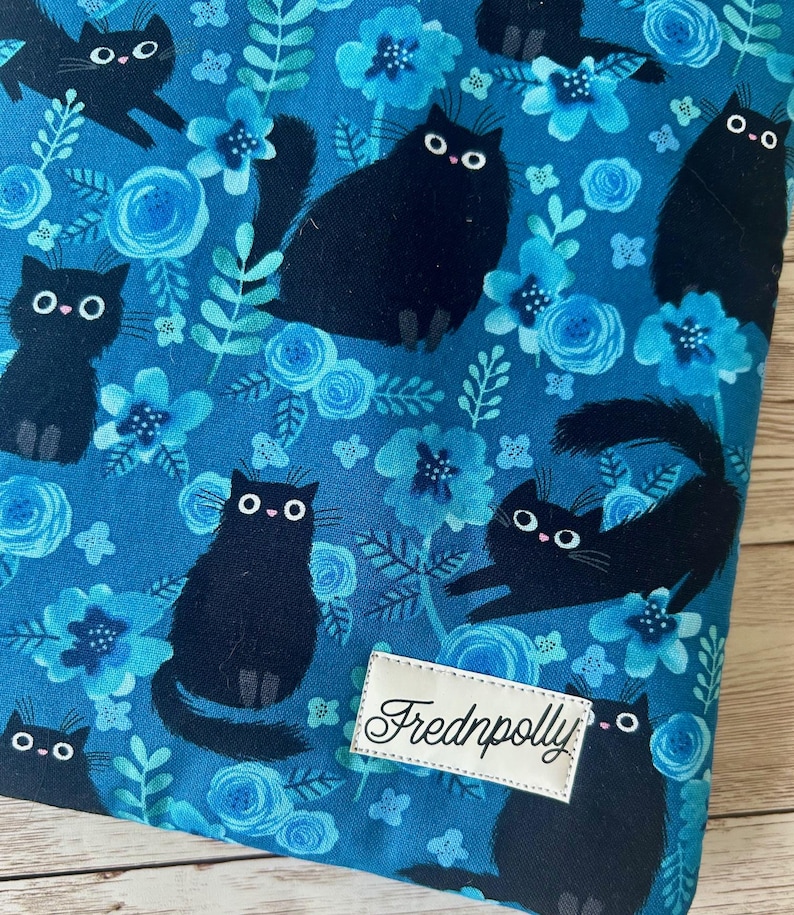 Puede incluir: Tela azul con un patr&oacute;n repetido de gatos negros y flores azules. La tela tiene una etiqueta blanca con el texto "Frednpolly" en ella.