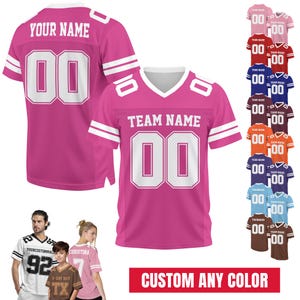 Puede incluir: Una camiseta deportiva rosa con texto blanco que dice YOUR NAME y TEAM NAME con el número 00 en blanco en la parte delantera y trasera. La camiseta tiene rayas blancas en las mangas y cuello en V. Se muestra una paleta de colores de varias opciones.