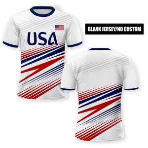 Puede incluir: Camiseta de f&uacute;tbol blanca con un dise&ntilde;o a rayas rojo, blanco y azul. La camiseta tiene un cuello azul y una peque&ntilde;a bandera estadounidense en el pecho. El texto "USA" est&aacute; impreso en azul en la parte delantera de la camiseta. CAMISETA EN BLANCO/SIN PERSONALIZACI&Oacute;N.