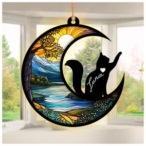 May include: A stained glass style ornament featuring a crescent moon with a black cat silhouette inside. The moon is set against a colorful landscape with a river, mountains, and a tree. The cat has a white heart on its chest and the name "Luna" written underneath.