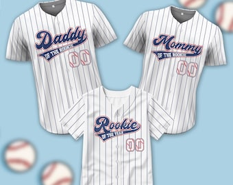 May include: Three white baseball jerseys with blue stripes. The jerseys have the text "Daddy of the Rookie", "Mommy of the Rookie", and "Rookie of the Year" on them. Each jersey has a number "00" on the front.