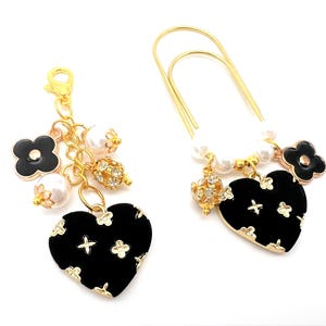 May include: A gold-toned lobster clasp charm and a large gold-toned paperclip, both featuring black heart-shaped pendants with gold floral accents, white faux pearls, and small gold-toned beads with clear crystals attached to the metal links.