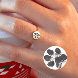 May include: A gold ring featuring a paw print design. The ring is worn on a person's finger. A black and white paw print sticker is on the person's hand.