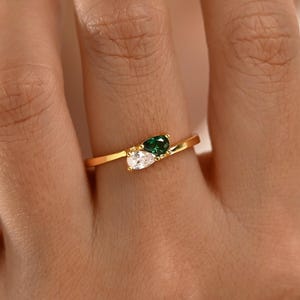 Puede incluir: Un anillo de oro con diseño de bypass y dos piedras en forma de pera. Una piedra es una gema blanca transparente y la otra es una gema de color verde intenso. La banda es fina y pulida, mostrando una estética sencilla con las dos piedras juntas.