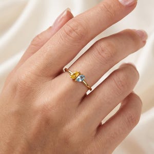 May include: A gold ring featuring two pear-shaped gemstones set side-by-side. One stone is a vibrant yellow, and the other is a light blue. The band is thin and polished, showcasing a minimalist design that highlights the contrasting colours of the stones.