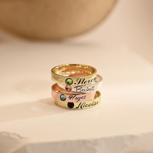 May include: Three personalised rings stacked on a white surface. The top ring is gold with the name "Steven" and a green gemstone. The middle ring is rose gold with the name "Beckett" and a pink gemstone. The bottom ring is gold with the name "Hayes" and a blue gemstone.