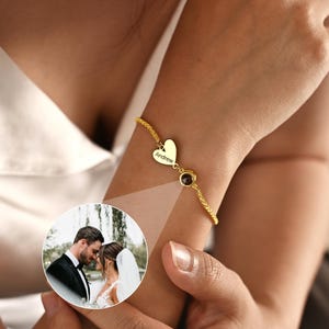 May include: A gold-toned bracelet featuring a delicate chain, a small heart-shaped charm engraved with the name Andrew, and a circular projection pendant that displays a photograph of a bride and groom in wedding attire.