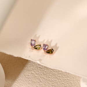 Peut inclure: Boucles d'oreilles en or rose avec trois pierres précieuses en forme de goutte, violettes et vert olive. Présentées sur une surface texturée blanc cassé.