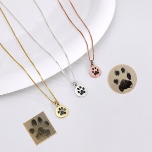 May include: Three necklaces with paw print pendants in gold, silver, and rose gold. Each necklace has a delicate chain. Two paw print impressions are also shown, one round and one square.