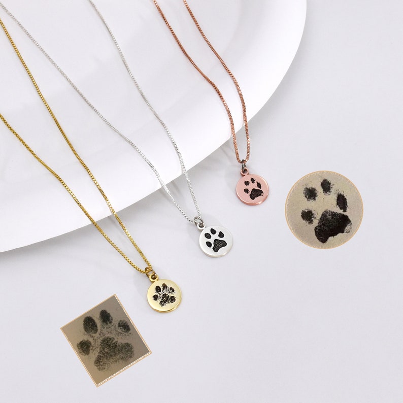 May include: Three necklaces with paw print pendants in gold, silver, and rose gold. Each necklace has a delicate chain. Two paw print impressions are also shown, one round and one square.