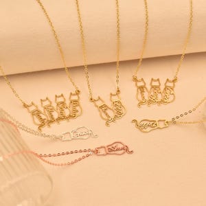 May include: Three gold necklaces with cat-shaped pendants. Each pendant is a silhouette of a cat sitting down, with the cat's name written inside the silhouette. The necklaces are on a light beige background.