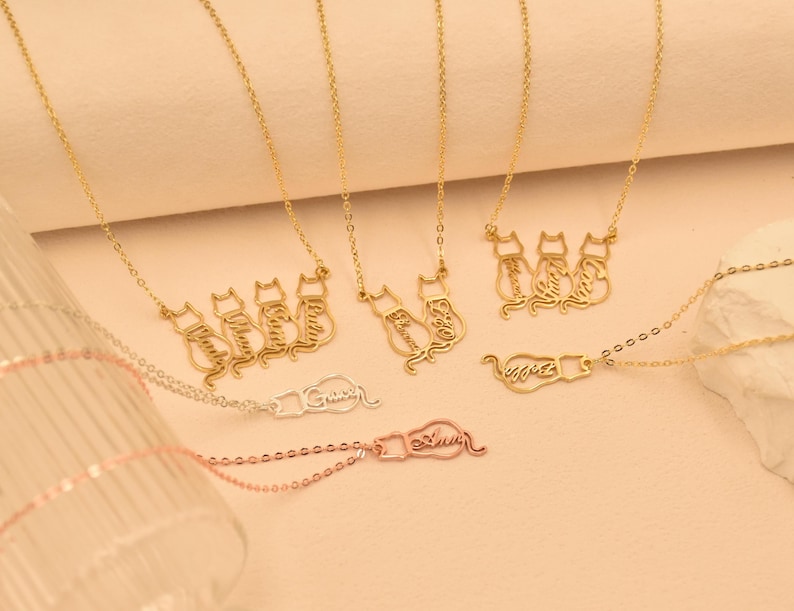 May include: Three gold necklaces with cat-shaped pendants. Each pendant is a silhouette of a cat sitting down, with the cat's name written inside the silhouette. The necklaces are on a light beige background.