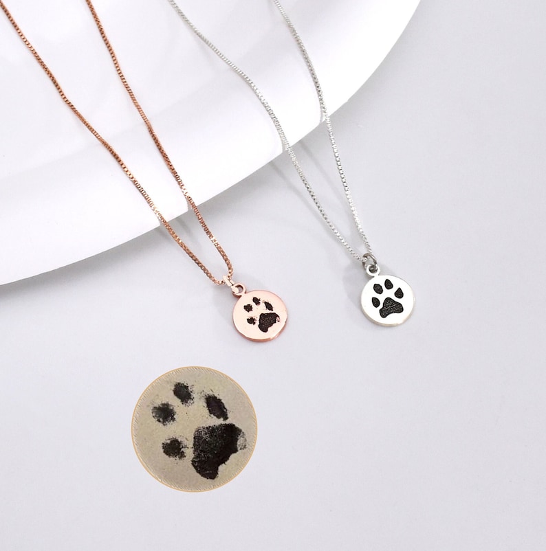 May include: Two delicate necklaces with round pendants featuring a paw print design. One necklace is rose gold, the other is silver. A third, larger, round pendant with a paw print is also shown. The necklaces are displayed on a white surface.