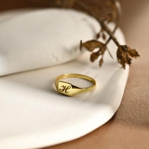 Può includere: Un anello sigillo dorato con finitura lucida e una faccia piatta rettangolare incisa con la lettera H in corsivo. La fascia è liscia e si restringe leggermente verso il retro, presentando un design classico con una lucentezza metallica.