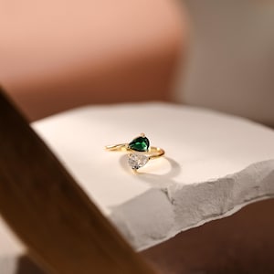 May include: A gold-toned open ring featuring two pear-shaped stones. One stone is a deep emerald green, and the other is a clear, colourless gemstone. The band is thin and polished, with the two stones positioned at the open ends of the ring.