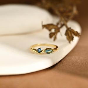 May include: A gold ring featuring a bypass-style band with two gemstones. One stone is a small, round, deep blue gem, and the other is a marquise-cut, teal-colored gem. The band has a smooth, polished finish and a gentle, flowing wave design.