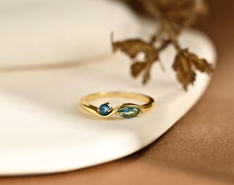 May include: A gold ring featuring a bypass-style band with two gemstones. One stone is a small, round, deep blue gem, and the other is a marquise-cut, teal-colored gem. The band has a smooth, polished finish and a gentle, flowing wave design.