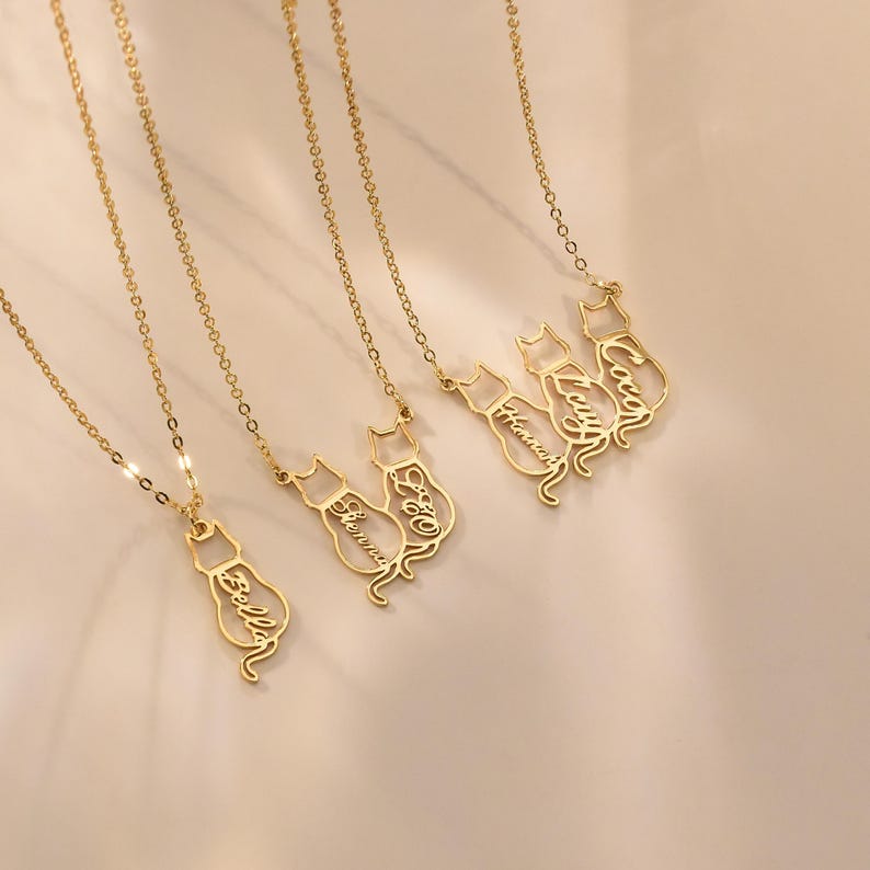May include: Five gold necklaces with cat-shaped pendants. Each pendant has a different name inscribed on it: Bella, Sienna, Leo, Lucy, and Coco.