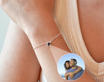 Puede incluir: Un brazalete de cadena de plata con un pequeño colgante de piedra roja oscura. El brazalete se lleva en la muñeca.