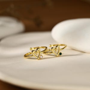May include: Two gold-toned open rings featuring a heart-shaped design. One ring is accented with a small, clear, round gemstone, while the other ring is accented with a small, dark green, round gemstone. Both rings have a smooth, polished finish.