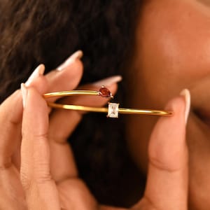 May include: A gold-colored bracelet with two gemstones. One gemstone is a pear-shaped, dark red stone, and the other is a rectangular, clear stone. The bracelet is held by a person with long nails.