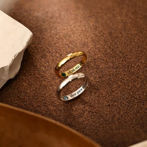 May include: Two rings, one gold and one silver, are shown. The gold ring has the engraved date "2026.02.03" and a green gemstone. The silver ring has the engraved phrase "Vivir para mi" and a blue gemstone.