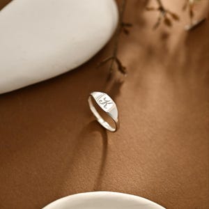 以下が含まれることがあります： 光沢のある金属仕上げのシルバーシグネットリング。平らな長方形の面には筆記体の「K」が刻印されています。リングは滑らかで先細りのバンドが中央のプレートにつながっており、反射するきれいな表面が特徴です。