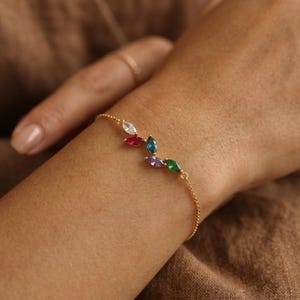 Könnte beinhalten: Ein zartes goldenes Armband mit einer Reihe farbiger, marquiseförmiger Edelsteine. Die Edelsteine sind in einem blattartigen Muster angeordnet, mit Farben wie Weiß, Rot, Blau, Lila und Grün. Das Armband wird am Handgelenk getragen.