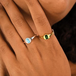 Könnte beinhalten: Zwei Siegelringe, einer aus Silber und einer aus Gold, jeweils mit einem rechteckigen Edelstein. Der Silberring hat einen hellblauen Stein, während der Goldring einen grünen Stein aufweist. Die Ringe werden an einer Hand gezeigt.