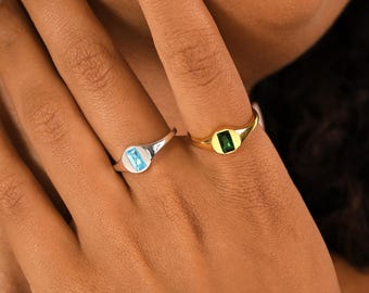 May include: Two signet rings, one silver and one gold, each featuring a rectangular gemstone. The silver ring has a light blue stone, while the gold ring showcases a green stone. The rings are displayed on a hand.