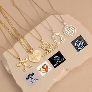 Puede incluir: Una colección de collares dorados y plateados con varios diseños de colgantes. Los colgantes incluyen un corazón con un perro, tijeras, una nube geométrica con la palabra "KOMPUTE" y formas abstractas. Los collares se muestran sobre una superficie beige claro.