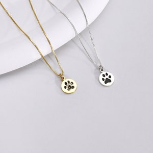 May include: Two delicate necklaces with round pendants featuring paw print designs. One necklace is gold-toned, the other is silver-toned. Both have fine chain links and are displayed on a light gray surface.