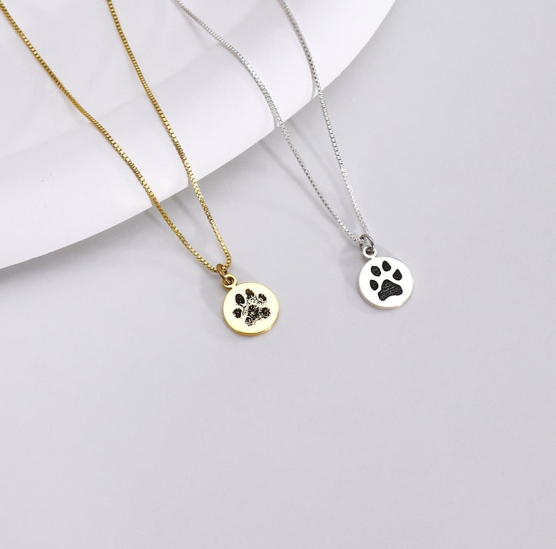 May include: Two delicate necklaces with round pendants featuring paw print designs. One necklace is gold-toned, the other is silver-toned. Both have fine chain links and are displayed on a light gray surface.